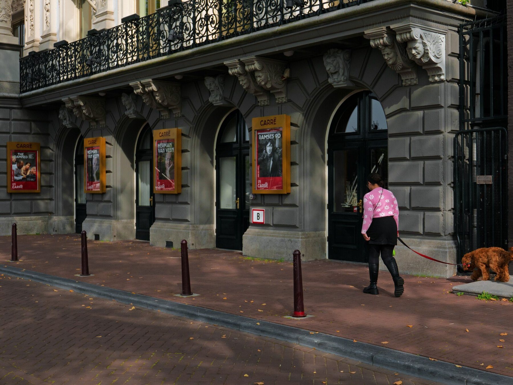 The arched facade of the Carré theatre in Amsterdam; a woman walks a brown dog along the brick pavement.